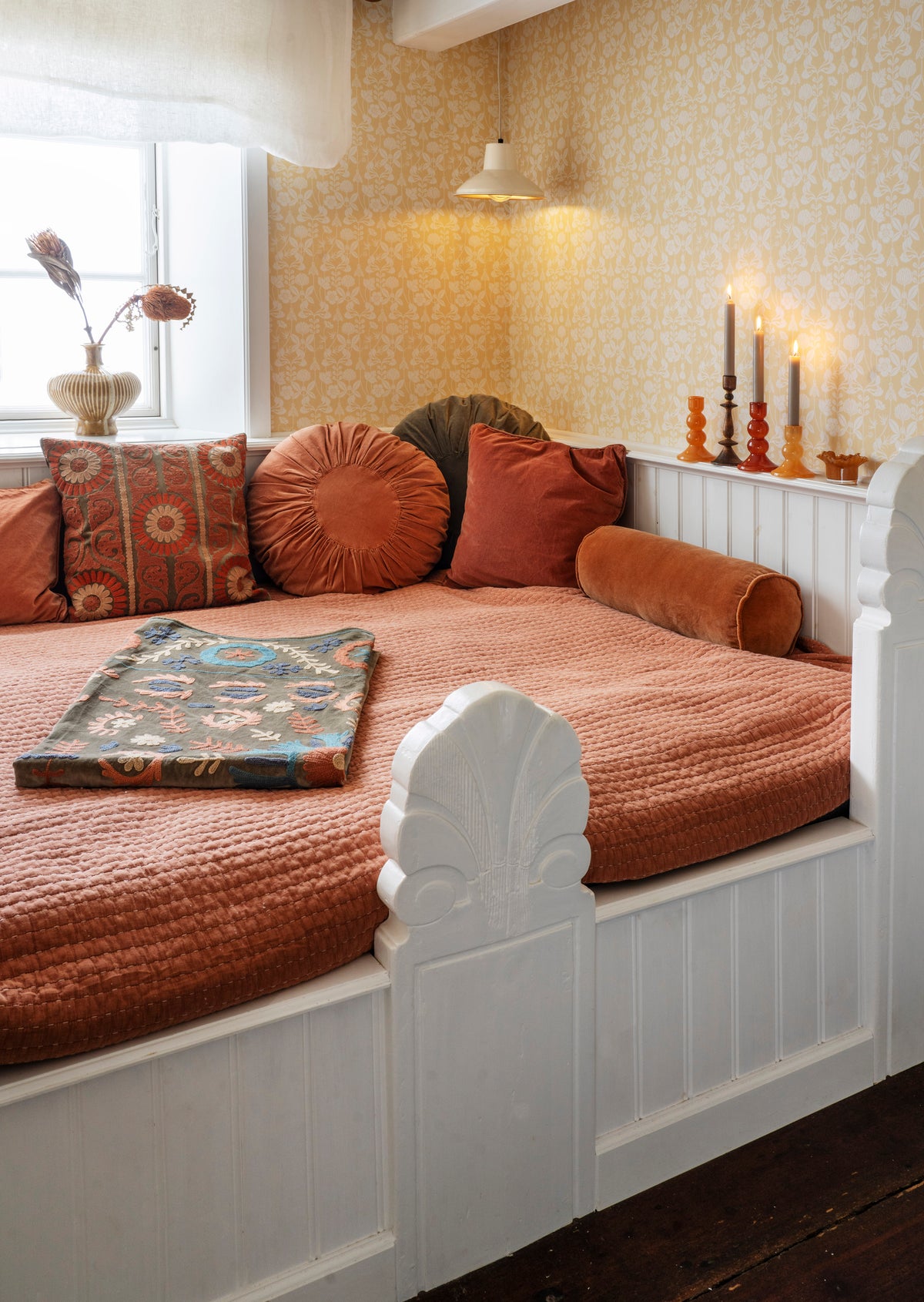 Rust orange Madam Stoltz Round Velvet Cushion on a daybed, surrounded by other cushions, a quilted throw, and patterned wallpaper.