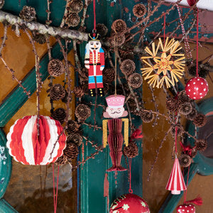 Hand-Painted Christmas Soldier by Madam Stoltz hangs with a straw star and paper ornaments on a festive branch adorned with pine cones.