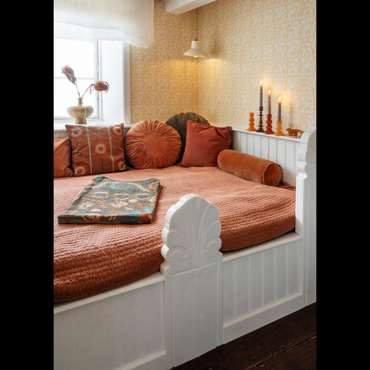 Cozy daybed featuring a Madam Stoltz Round Velvet Cushion among various pillows, orange quilt, and patterned wallpaper.