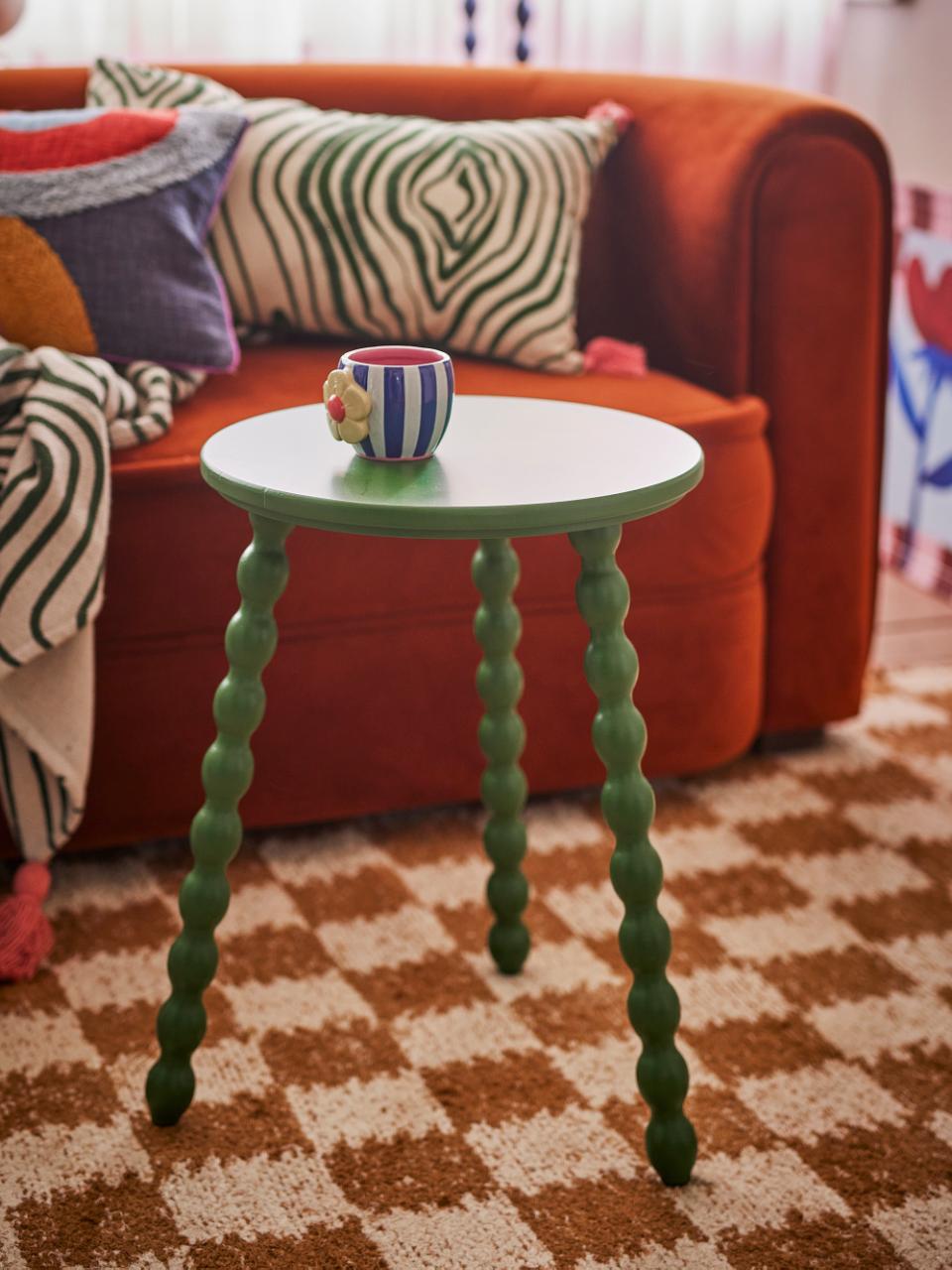 Bloomingville Salino Side Table with green bobble legs, holding a striped cup, next to an orange sofa with patterned pillows.
