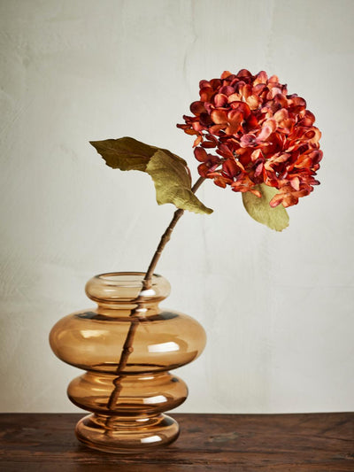 Bloomingville artificial hydrangea stem with vibrant reddish-orange flowers and green leaves, in a brown glass vase.