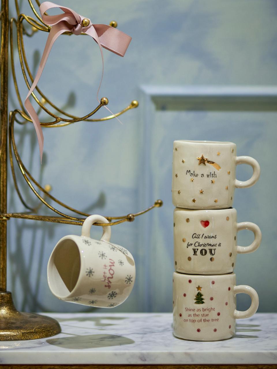 Christy Cup Multipack, Set of 4 by Bloomingville. Cream festive mugs with holiday phrases &amp; designs, displayed on a gold tree.