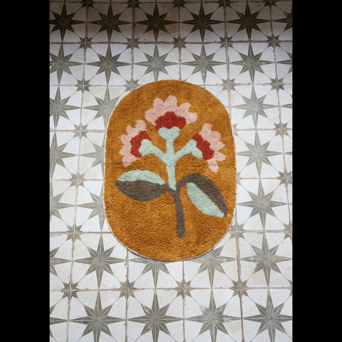 Madam Stoltz oval tufted bath mat in mustard yellow with a pink, red, and green floral design on a star-patterned tiled floor.