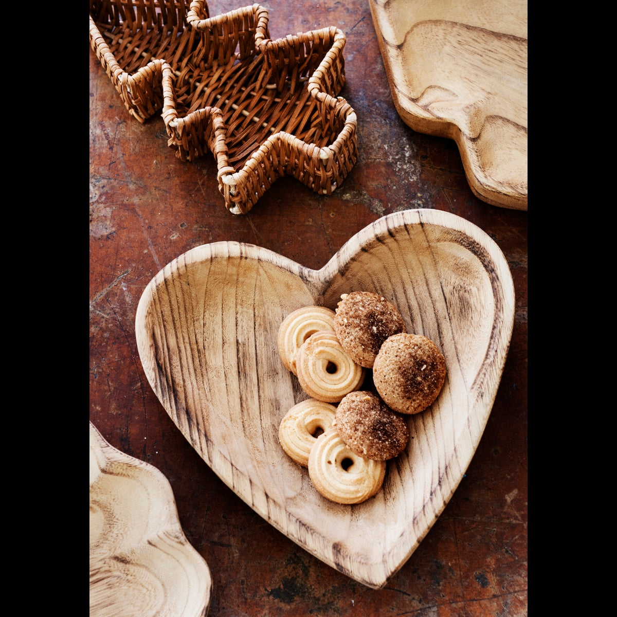 Madam Stoltz wooden heart tray filled with cookies, surrounded by other decorative wooden and wicker trays.
