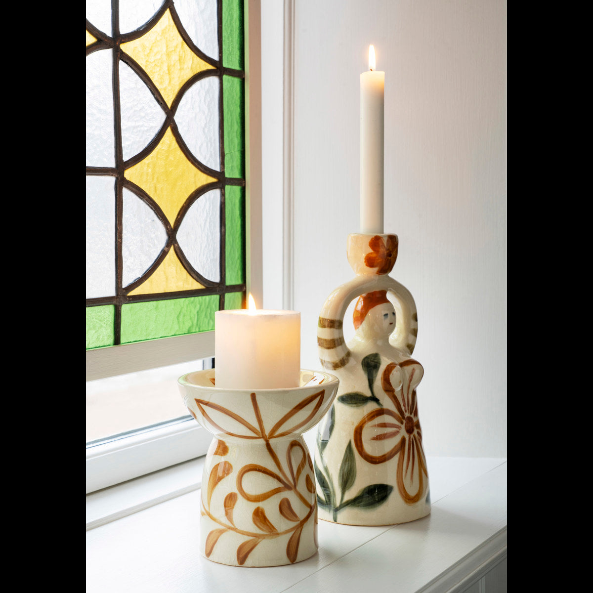 Hand-Painted Candle Holder Figurine by Madam Stoltz and a decorated pillar holder with lit candles on a sunny windowsill.