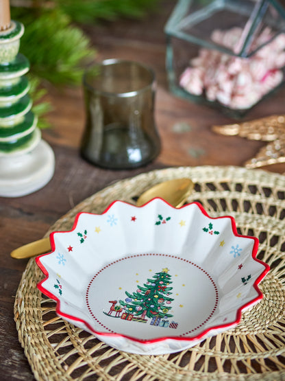 Bloomingville Naive Bowl, Red, a white festive bowl with a Christmas tree, gifts, snowflakes, and holly pattern, on a woven placemat.