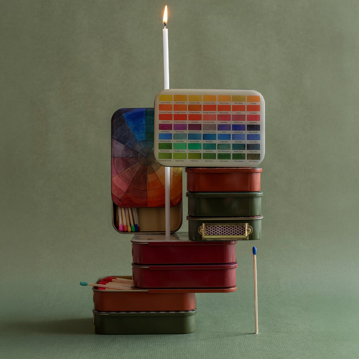 Stacked Roomytown Matches in Tin with a lit candle, color swatch, and colorful matchsticks on a green surface.