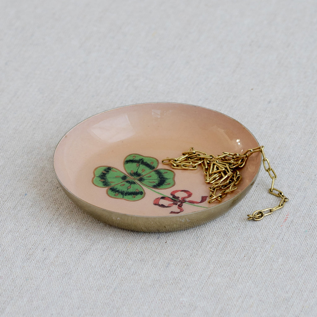 Roomytown Round Enamel Tray in peach, featuring a four-leaf clover and red bow design, holding a gold chain.