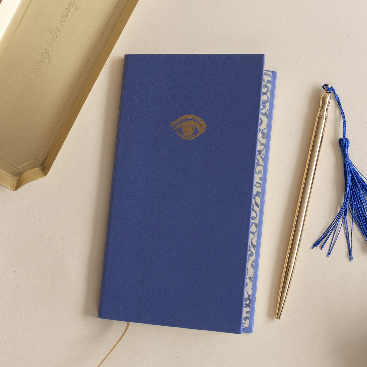 Slim Lined Notebook from Roomytown in blue with a gold eye design, alongside a gold pen with a blue tassel.