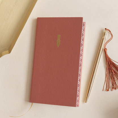 Roomytown Slim Lined Notebook in coral with a golden pen icon, next to a golden pen with a rust tassel, and a &